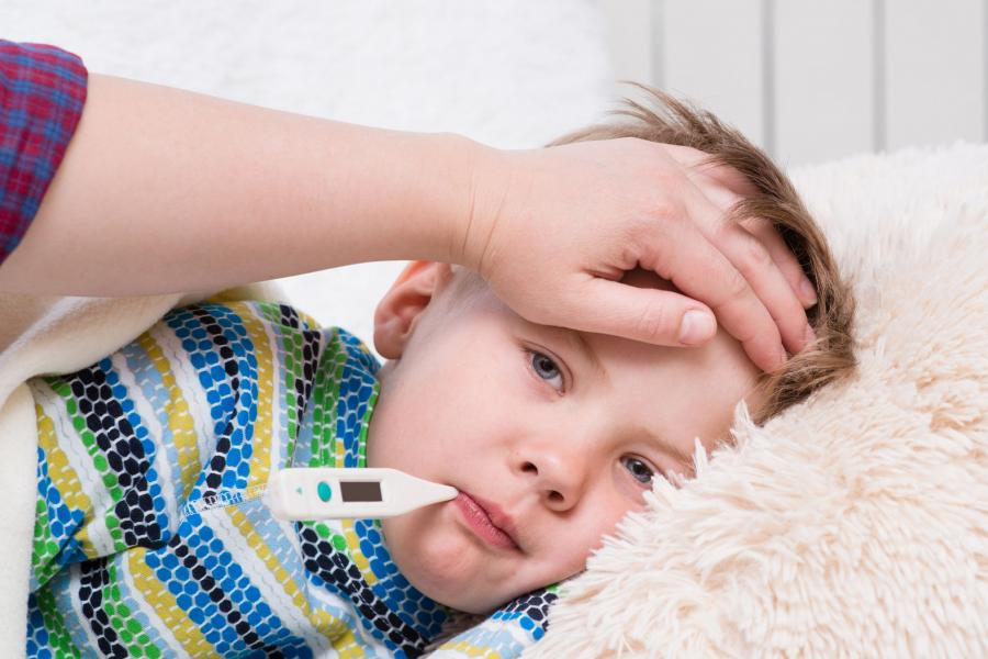 Sick young boy lying in bed with a thermometer in his mouth and his mother's hand on his forehead