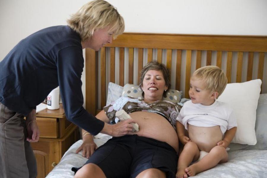 A pregnant mother lying on her bed. Her young child is looking on as the lead maternity carer examines the mother's tummy