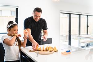 A father chopping up fruit in the kitchen while his daughter eats it and looks on