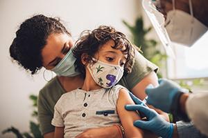 A boy wearing face mask having his COVID-19 vaccine while sitting on his mum's lapat home.